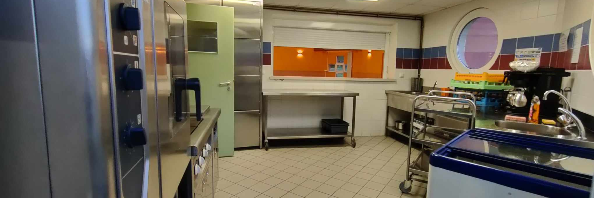 Cuisine professionnelle avec réfrigérateur, évier, lave-vaisselle, chariots et plans de travail.