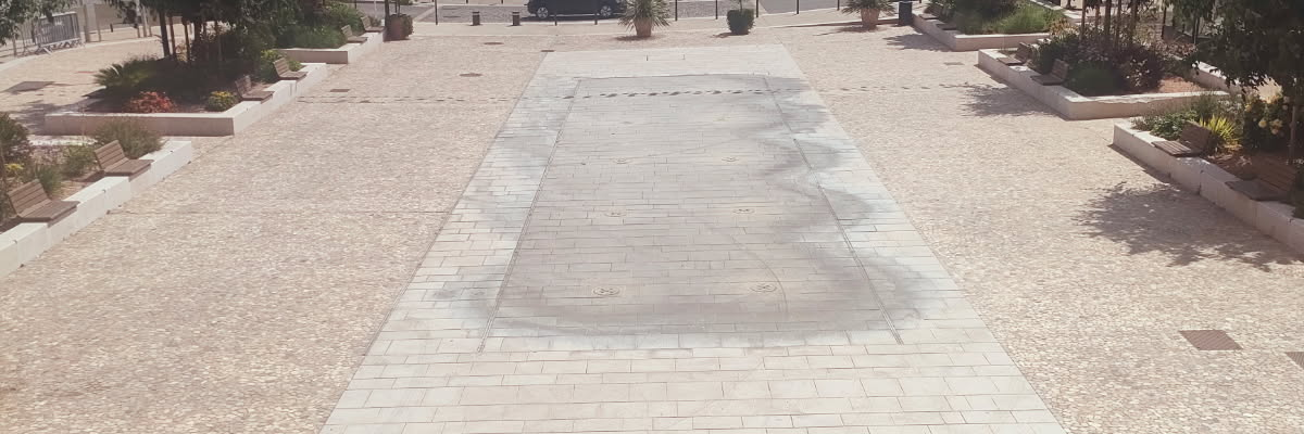 Place pavée avec bâtiments, arbres, barrières et pots de fleurs.