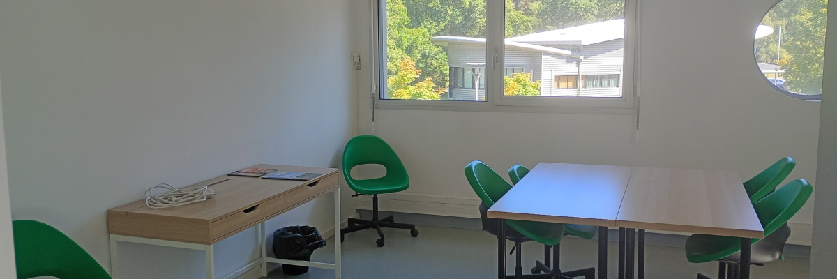 Salle avec tables, chaises vertes, étagère, fenêtre et porte ronde.