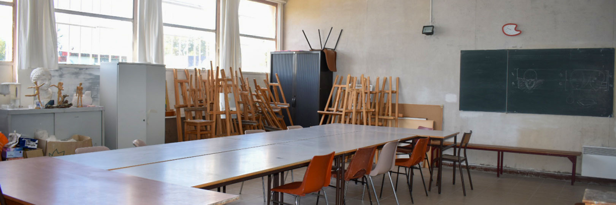 Salle de classe avec tableau noir, tables, chaises, fenêtres et armoire.