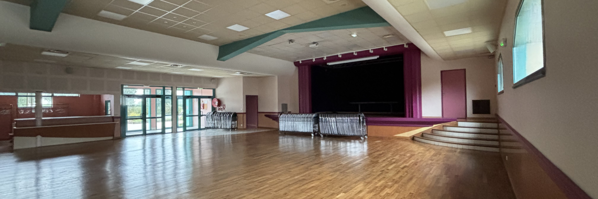 Salle vide avec plancher en bois, grandes fenêtres, scène, escalier et éclairage au plafond.