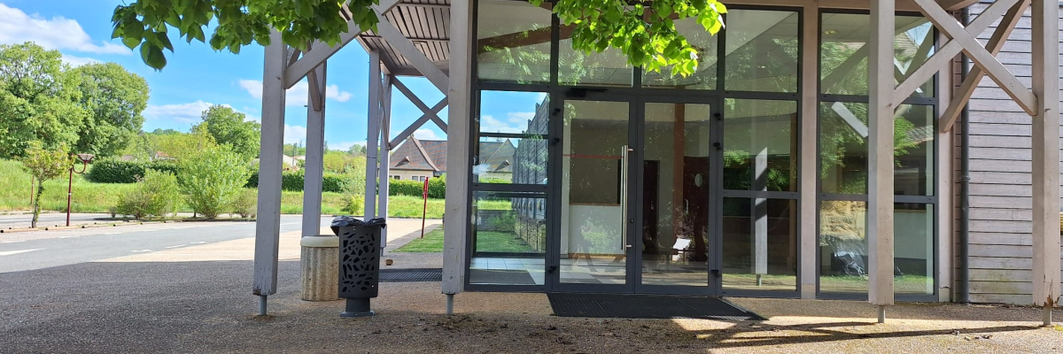 Bâtiment avec grandes fenêtres, porte vitrée, arbre, pelouse, allée et poubelle.