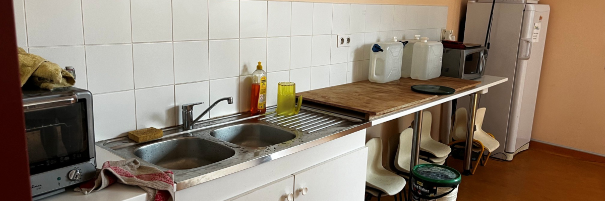 Cuisine avec évier, micro-ondes, réfrigérateur, plan de travail, produits de nettoyage et poubelle.