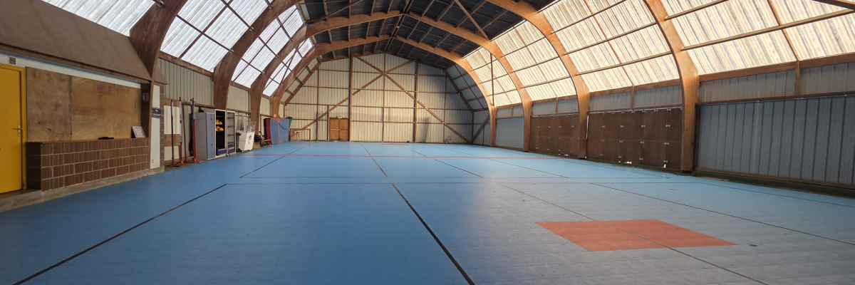 Gymnase avec sol bleu, toiture en arc, murs métalliques et portes en acier.