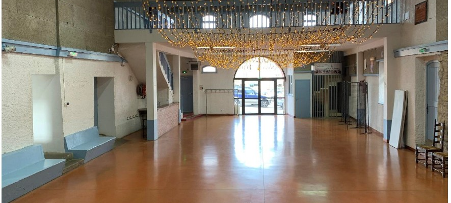 Grande salle vide avec scène, escalier, balustrade, éclairage suspendu et portes vitrées. Dimensions : salle 17 m x 10 m, scène 7 m x 3 m.