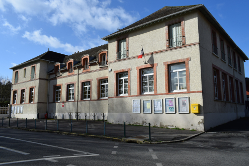 École avec murs beiges, fenêtres rouges, drapeaux, panneaux d'affichage et parking.
