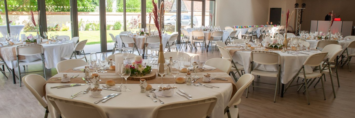 Salle de réception avec tables rondes, chaises, nappes, vaisselle et décoration florale.
