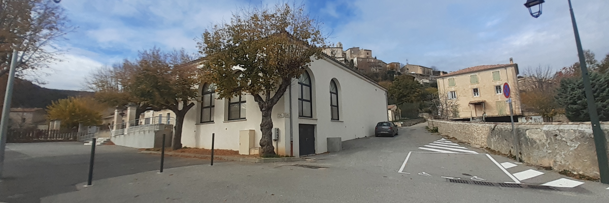 Rue avec bâtiments, arbres, lampadaires, passerelle piétonne et voitures.