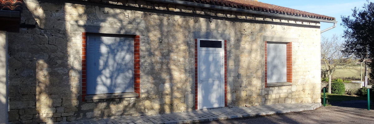 Bâtiment en pierre avec porte blanche et deux fenêtres aux volets rouges.