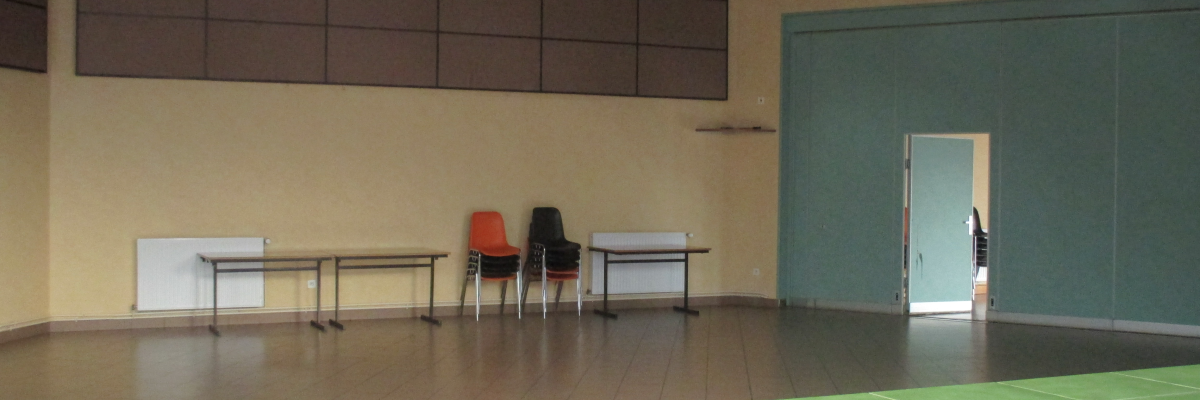 Salle vide avec tables, chaises, et tableau. Plante en premier plan et porte ouverte au fond.