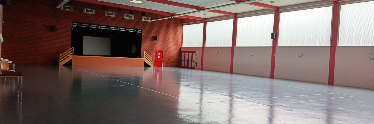 Salle spacieuse avec scène, sol lisse, murs rouges et blancs, grandes fenêtres et éclairage au plafond.