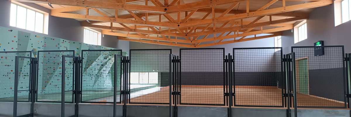 "Salle avec structure bois, tables, chaises, mur d'escalade vert et sol gris."