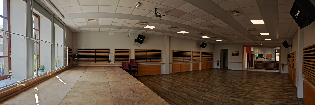 Salle spacieuse avec grandes fenêtres, rideaux, haut-parleurs suspendus, porte, extincteur et sol en bois.