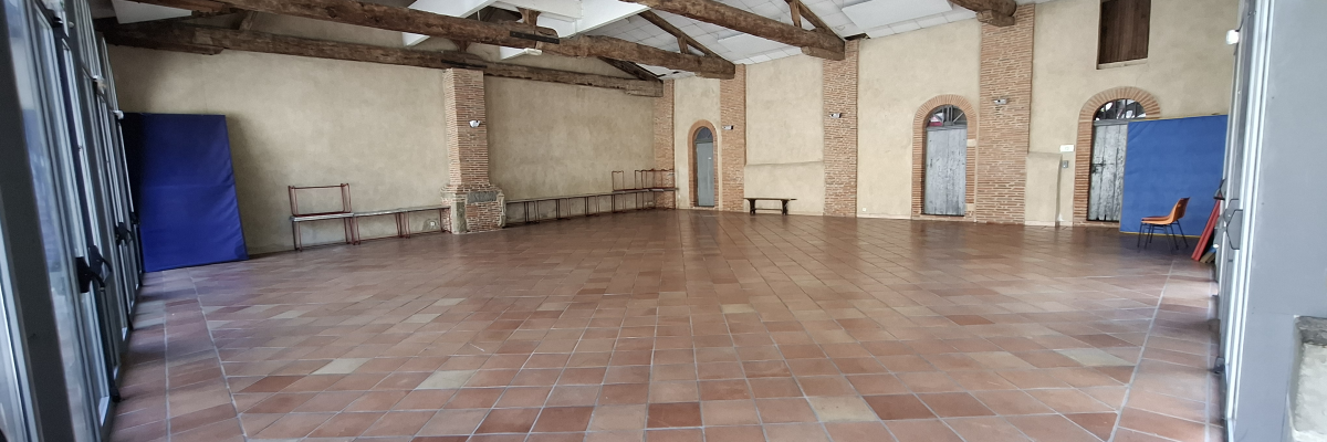 Salle spacieuse avec sol carrelé, poutres apparentes, bancs, portes et fenêtres.