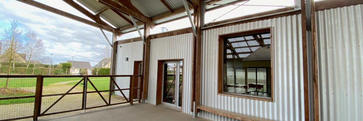 Atelier avec sol en béton, toit en tôle, poutrelles apparentes, porte et fenêtre.