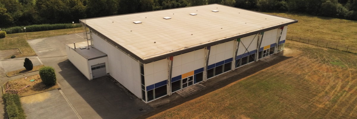 Bâtiment industriel avec toit plat, grandes fenêtres et porte de chargement, entouré de pelouse et de forêt.