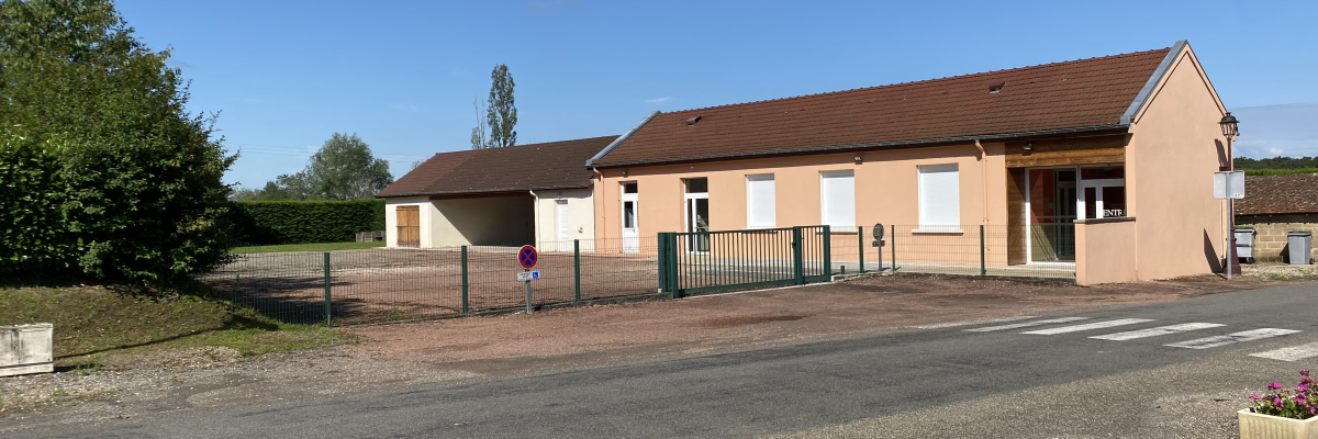 Bâtiment beige avec toit brun, clôture verte, porte blanche, et arbre à gauche.
