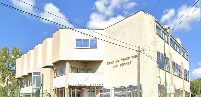 Batiment beige de deux étages avec de grandes fenêtres. Entrée principale avec escalier et porte en métal. Panneaux indiquant "Maison des Passeports" et "Jean Monnet". Ciel bleu avec quelques nuages.