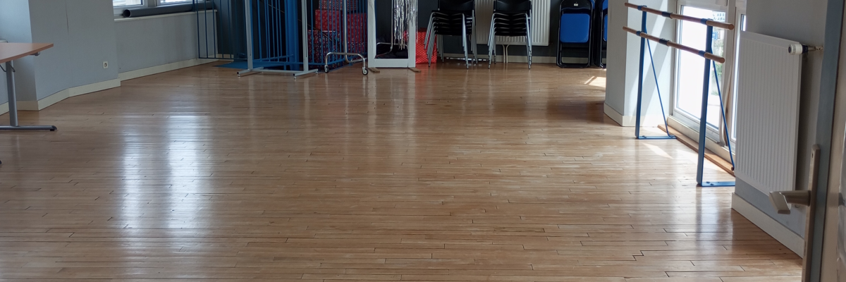 Salle vide avec parois vitrées, plancher en bois, barre d'appui, bancs, et matériel de sport.