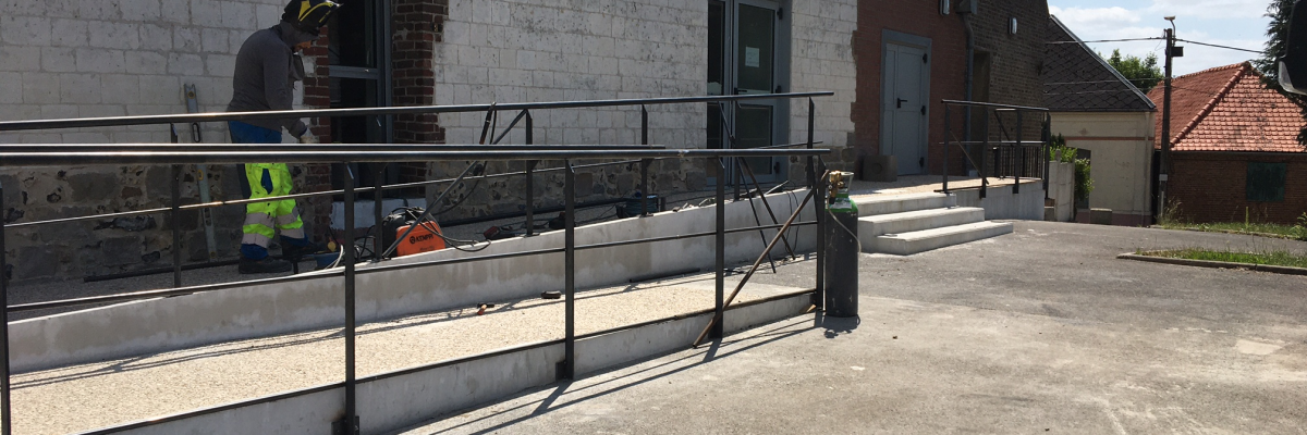 Bâtiment avec rampe d'accès et garde-corps métalliques. Deux personnes en gilet de sécurité près de l'entrée.