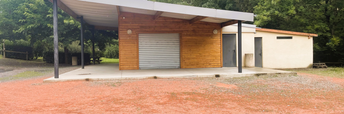 Un abri ouvert avec un toit en métal, des murs en bois et une porte de garage en métal. Derrière l'abri, un petit bâtiment blanc est visible. L'ensemble est situé dans une zone herbeuse avec des arbres en arrière-plan.