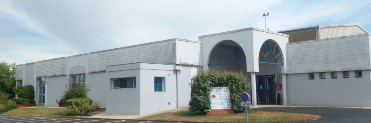 Bâtiment industriel avec façade blanche, arches d'entrée, parking et panneaux de signalisation.