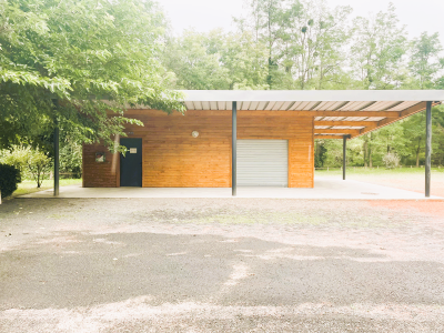 Petit bâtiment en bois avec porte noire, garage et auvent. Allée gravillonnée et végétation.