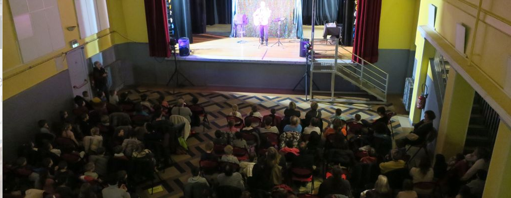 Salle de spectacle avec scène, rideaux, projecteurs et public assis.