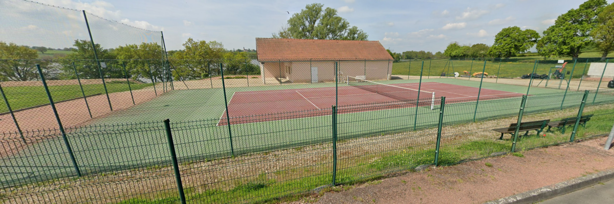 Terrain de tennis avec clôture, filet et banc. Bâtiment adjacent.