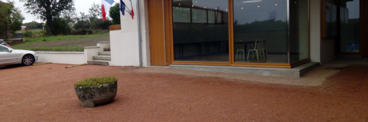 Bâtiment avec grandes fenêtres, porte en bois, drapeaux, pot de fleurs, voiture et banc.