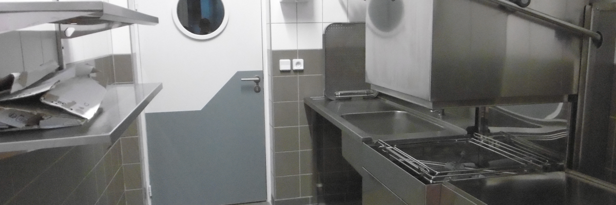 Cuisine industrielle avec plan de travail, fours, plaque de cuisson, étagère et porte.