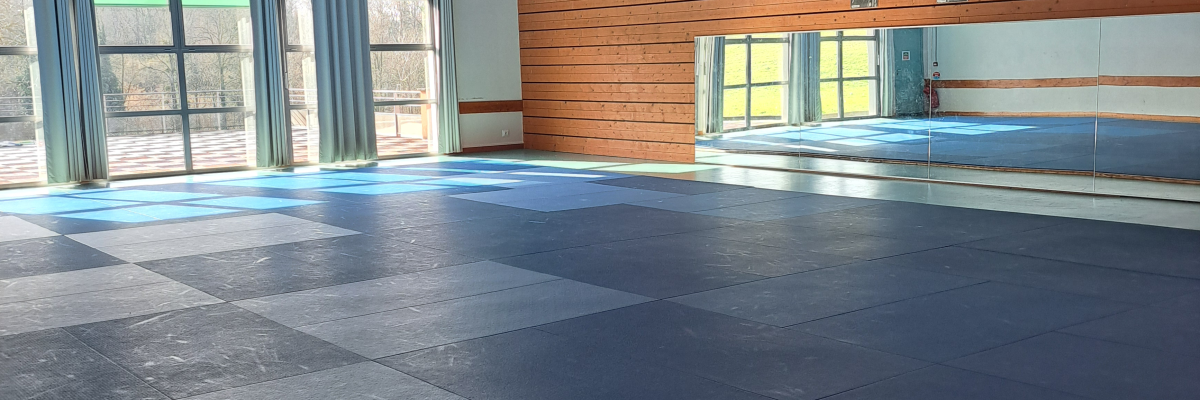 Salle avec sol bleu, poutres apparentes, fenêtres, rideaux et éclairage suspendu.