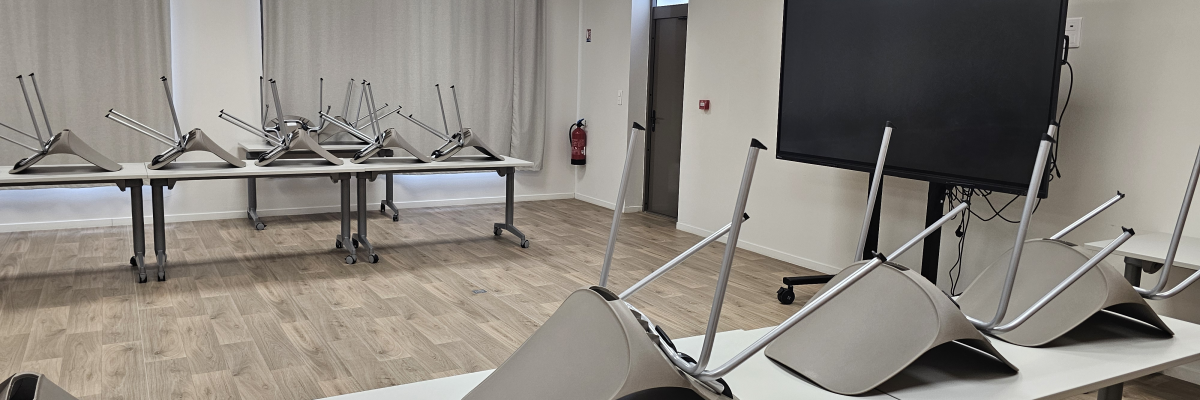Salle avec tables, chaises empilées, écran et tableau blanc.