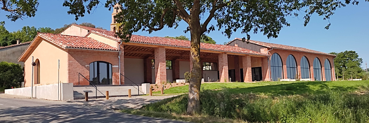 Bâtiment avec toit en tuiles, murs en brique, grandes fenêtres cintrées, arbre et pelouse.