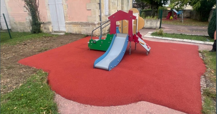 Aire de jeux avec toboggan, escalier et glissade, sur sol en caoutchouc rouge, près d'un bâtiment.