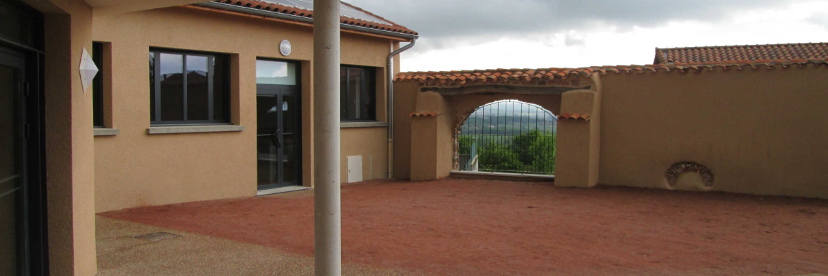 Bâtiment extérieure avec sol en gravier, murs beiges, fenêtres, porte vitrée, arbre et mur d'enceinte.