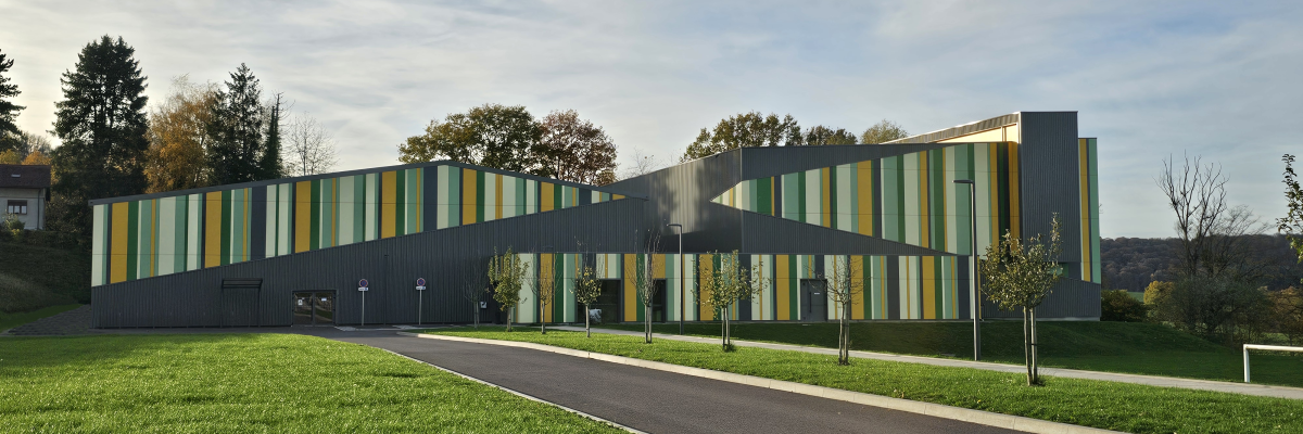 Bâtiment avec façade grise et jaune, allée, arbres, gazon.