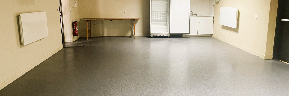 Pièce avec sol gris, radiateurs, plan de travail, extinguishateur, réfrigérateur et évier.