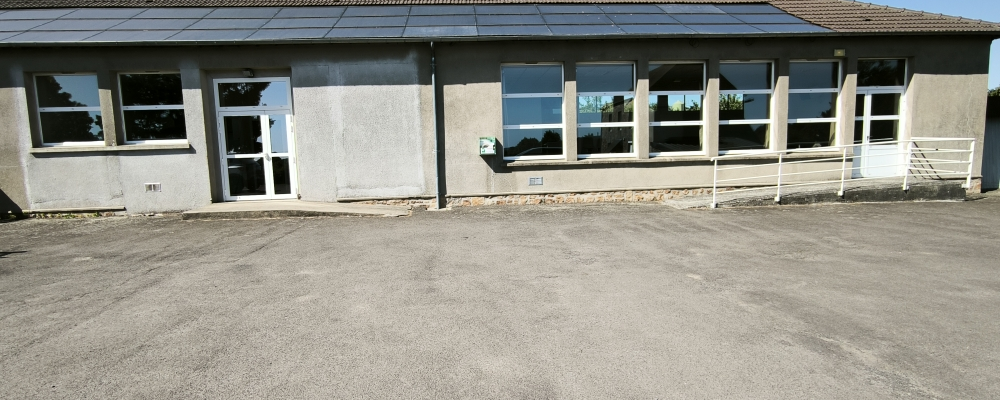 Bâtiment à un étage avec panneaux solaires sur le toit, entrées doubles vitrées, et porte d'entrée en verre. Parking en gravier devant.