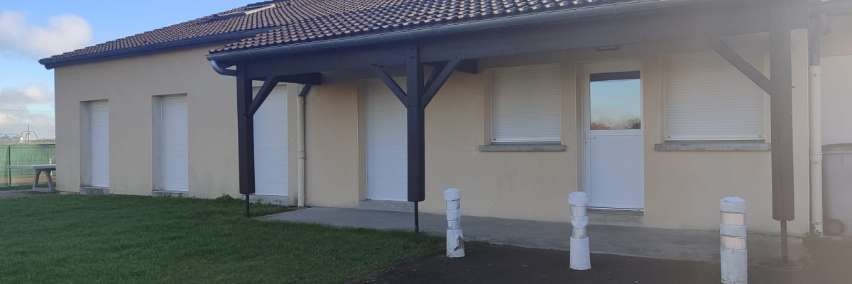 Bâtiment à toit en tuiles, murs crème, portes et fenêtres blanches. Poteaux blancs devant. Pelouse à gauche.