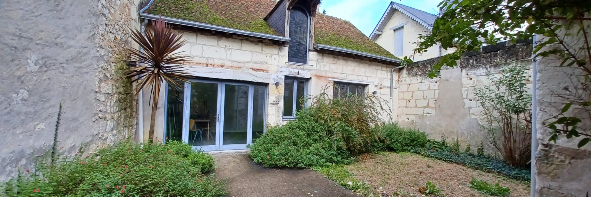 Maison avec jardin, mur en pierre, toit en tuiles, porte vitrée, buissons, plantes, allée.