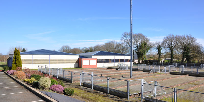 Terrain de sport avec clôture, projecteurs et parking adjacent.