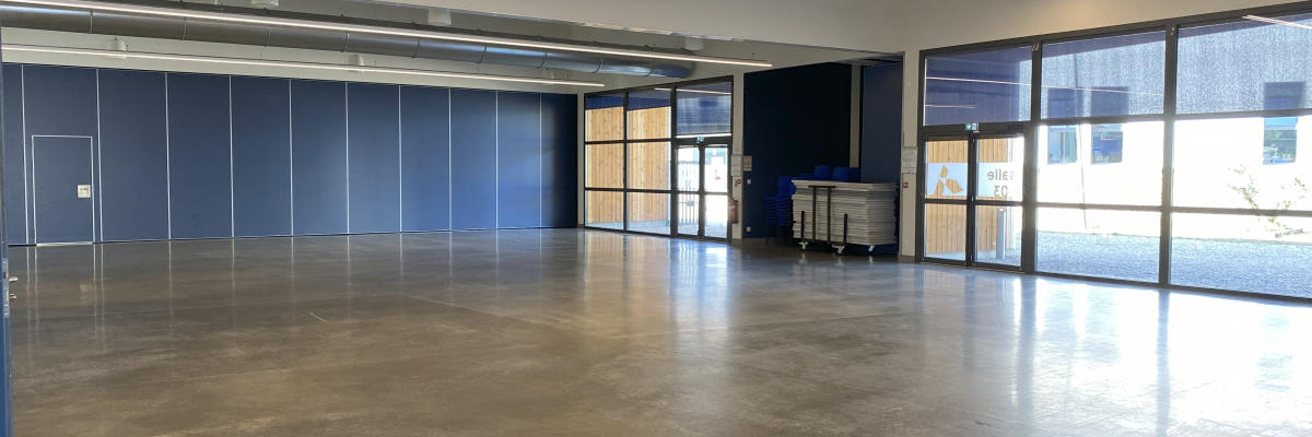 Grande salle vide avec sol en béton, grandes fenêtres, murs bleus et portiques suspendus au plafond.