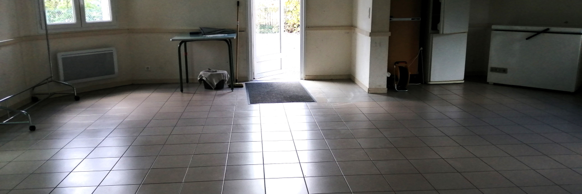 Salle avec sol carrelé, porte, fenêtre, radiateur, frigo, table pliable et balai.