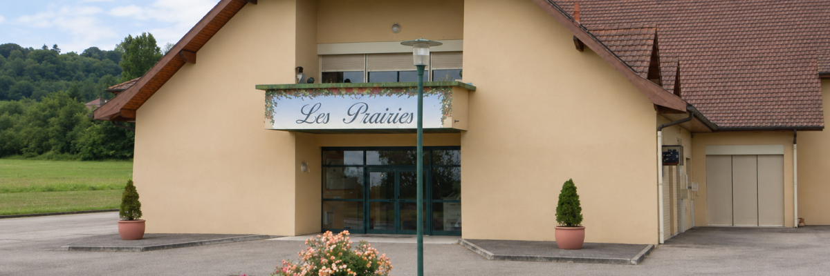 Bâtiment avec enseigne "Les Prairies", entrée vitrée, deux pots de fleurs, pelouse et parking.