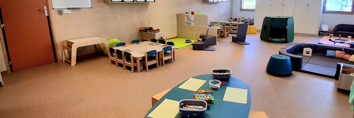Salle de classe avec table verte, chaises, bancs, table pour enfants, tableau blanc et armoires.