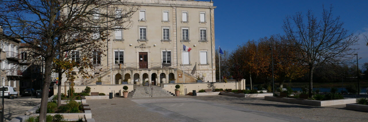 Place devant un bâtiment avec arbres, végétation et allée pavée.