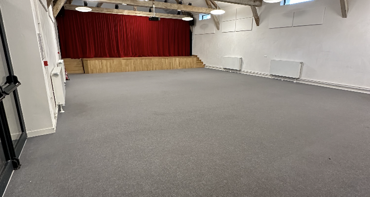 Salle vide avec poutrelles apparentes, sol gris, rideau rouge et grandes fenêtres.