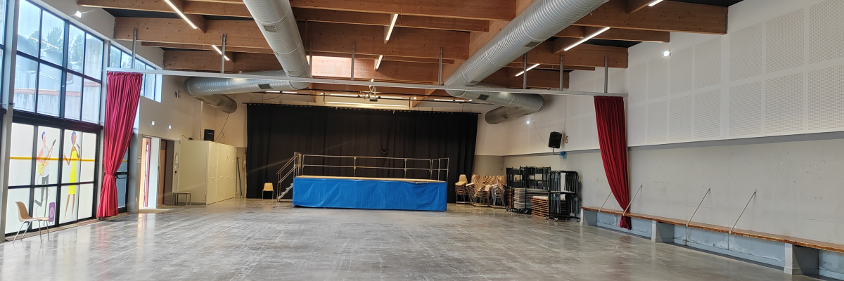 Salle avec sol en béton, murs blancs, plafond avec poutres et conduits, grande fenêtre, rideaux rouges, et divers équipements de théâtre.