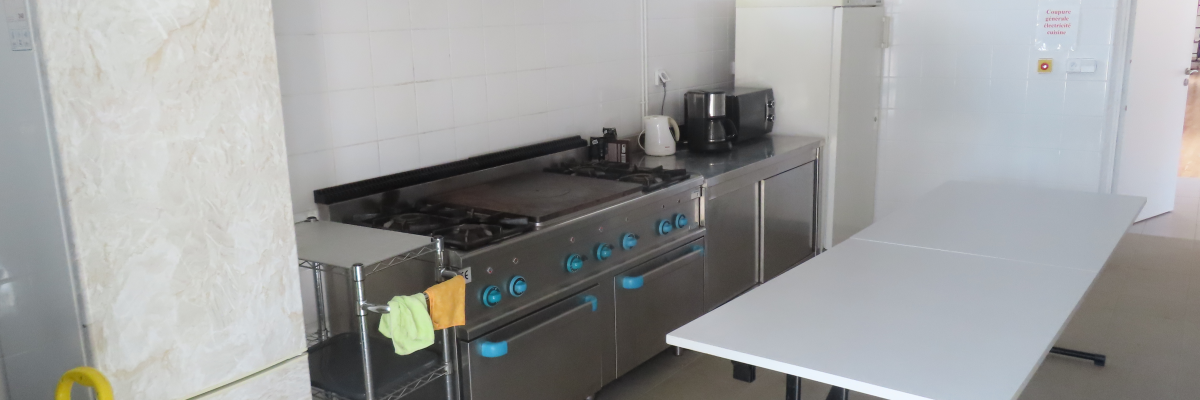 Cuisine avec plaque de cuisson, hotte, réfrigérateur, table et balai.